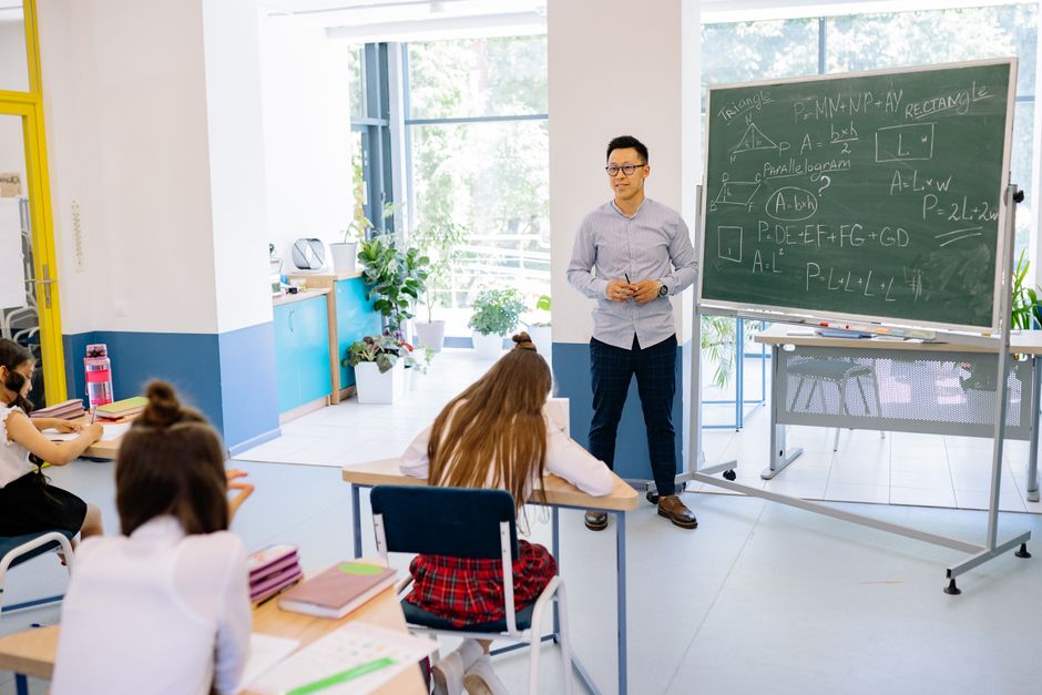 Nastavnik podučava učenike u svijetloj učionici s pločom ispunjenom matematičkim jednadžbama.